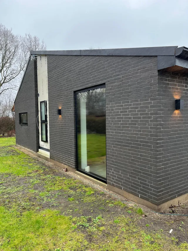 Tilbygning i mørke mursten med stort vinduesparti og udelamper